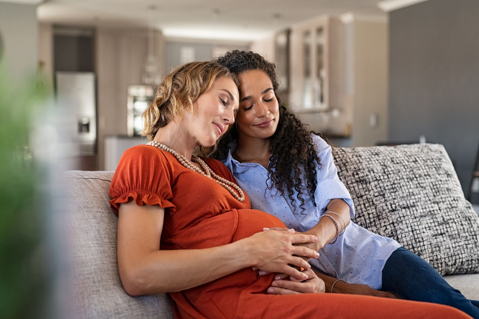 Two women cuddle on the couch, one with her hand on the other's pregnant belly