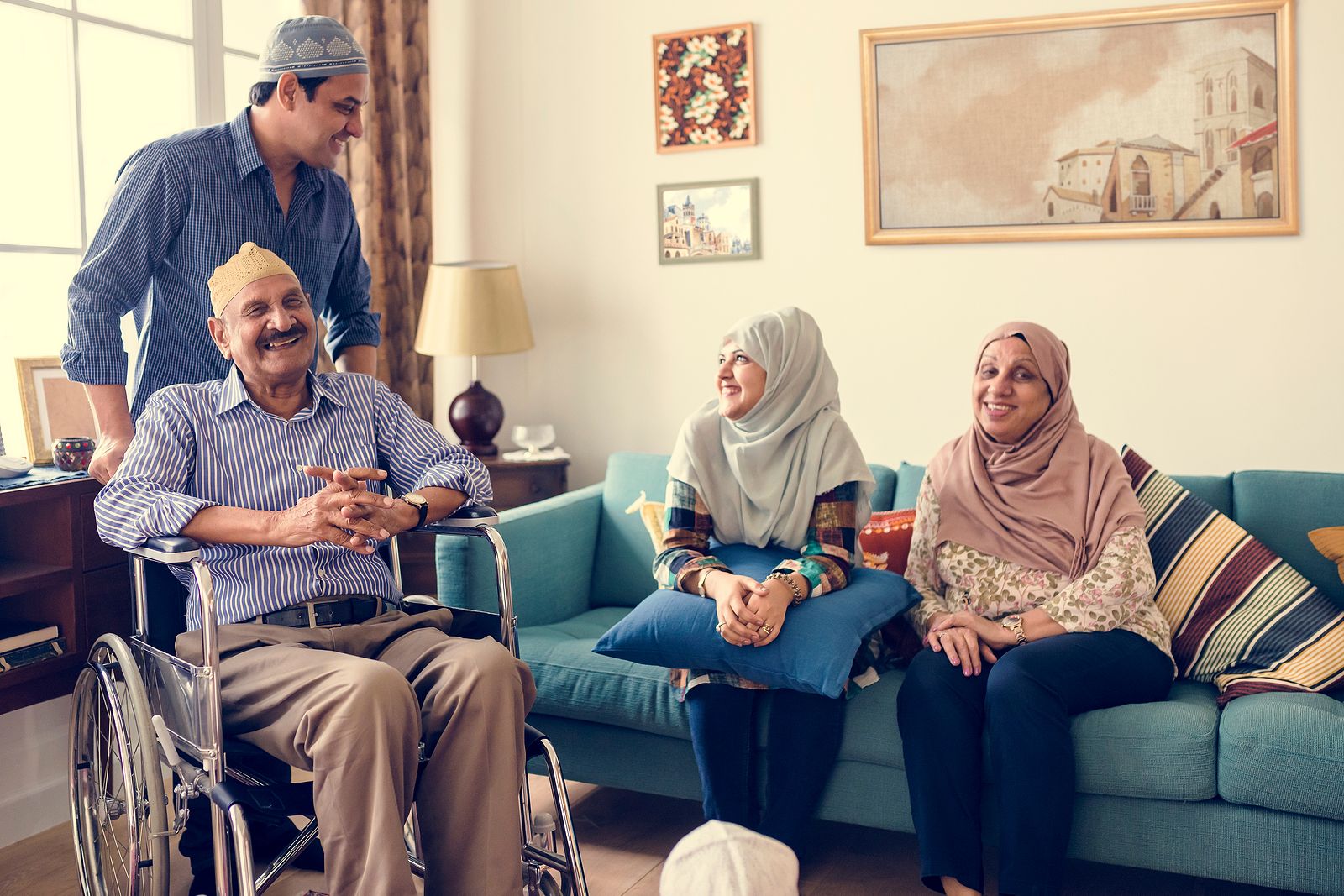 Middle Eastern family gathered in a room, laughing together as the older man is assisted in a wheelchair for medical care