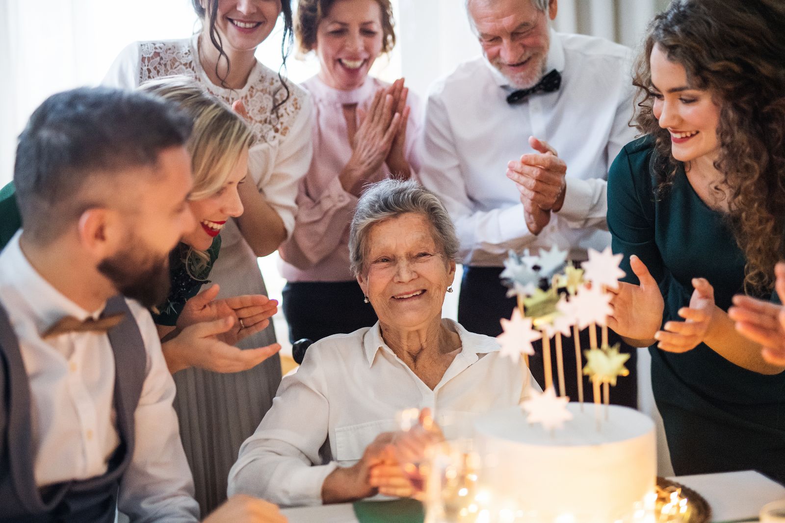 Smiling older woman surrounded by family and friends clapping as she sits in front of a lit birthday cake